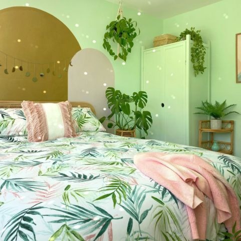 A green bedroom with lots of green plants in rattan baskets and planters. The ceiling light, rug and tray on the bed is also rattan. The bedding is a palm print. On the wall behind the bed is a set of three overlapping arches, with the middle one gold and the two either side a neutral tone.