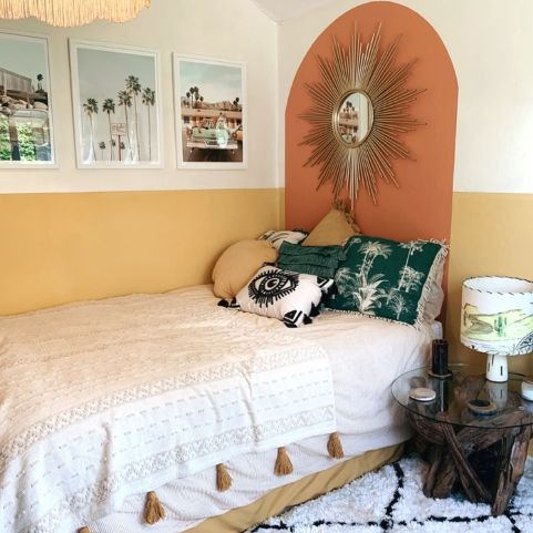 A boho style bedroom, with two tone walls. The bottom half of the wall is a gold colour and the top is an off white. There is an arch behind the bed in a rusty orange colour. On the arch there is a big sunburst mirror. On the bed there is a selection of coho cushions, including the furn. ashram eye cushion. There is a cream throw with ochre tassels on.