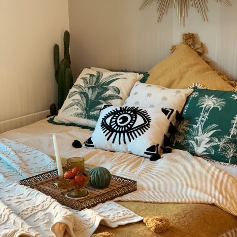 A boho style bed. Above the bed, we can see the bottom of a sunburst mirror. On the bed there is a selection of boho cushions, including the furn. ashram eye cushion. There is a cream throw with ochre tassels on. There is a cactus in the corner.