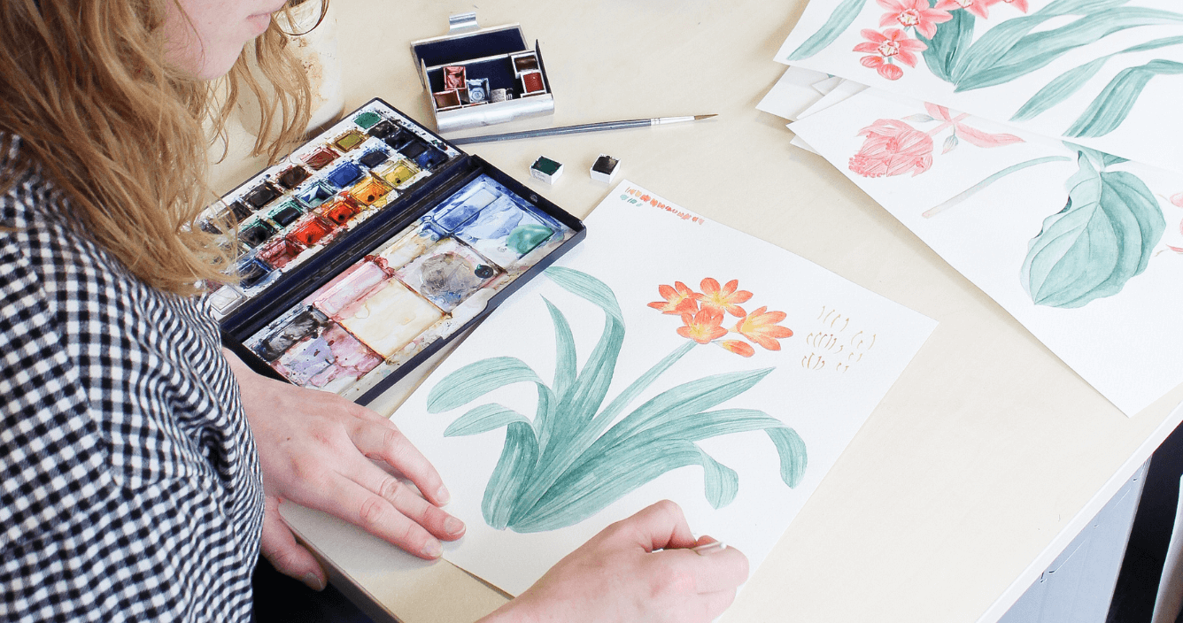 a desk with a watercolour picture of an orange flower with lots of leaves, as well as a watercolour paint palette, and a young female designer working on the painting
