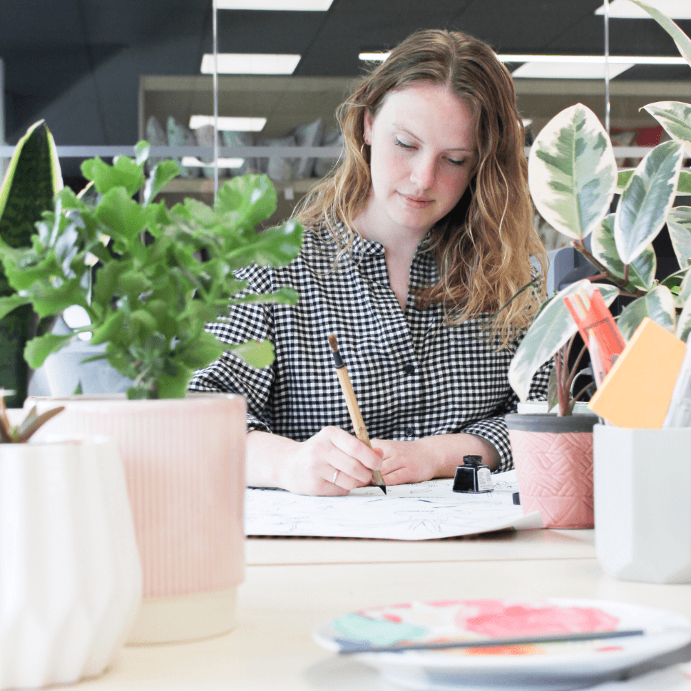 a young female designer draws at her desk. she is framed by plants. 