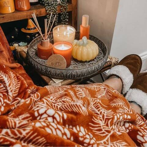 A rust orange fleece throw with an autumnal woodland design, placed over a person wearing slippers, next to a side table holding a home fragrance kit and some Halloween ornaments.