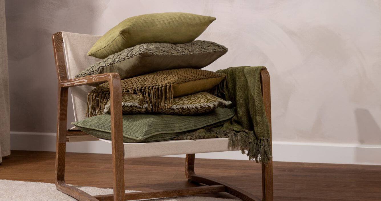 A selection of moss, olive and forest green cushions stacked on a wooden chair with a green throw draped over the arm, in a room with beige walls and a beige rug.