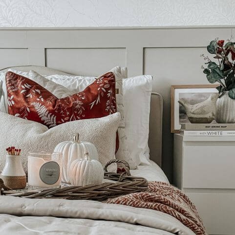 A cosy bed decorated with autumn home decor, including a white duvet cover, a woven rust throw, a velvet brick red cushion, a white bouclé cushion and a woven tray holding autumn trinkets.