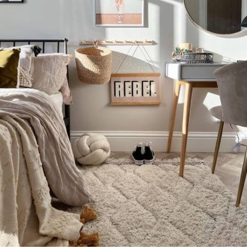 A bedroom decorated with cosy autumn home decor, including pale grey walls, a furry white rug, a bouclé knot cushion, bed cushions, throws and a desk.