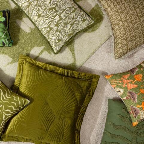 A selection of autumn themed green scatter cushions, including a quilted velvet cushion, floral and mushroom designs, scattered on a white surface with a coordinating green throw.