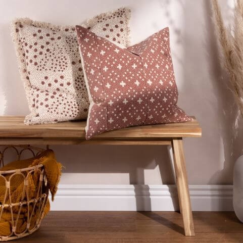 Two autumn style cotton cushions with abstract geometric and textured designs, displayed on a wooden bench next to a brown crinkle cotton throw in a storage basket.