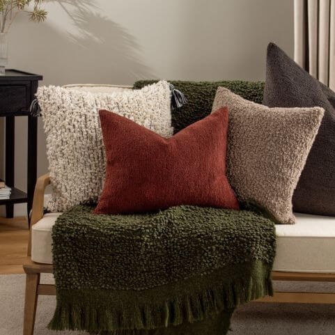 A selection of bouclé autumn home furnishings, including a white, red, brown and grey cushions, arranged on top of a green bouclé throw.