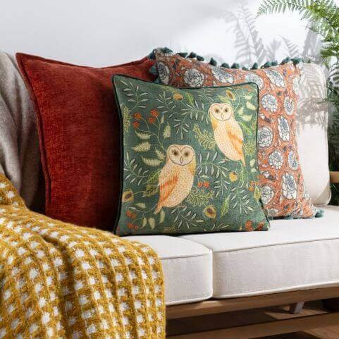 A selection of country style autumnal cushions, including a plain red cushion, a green cushion with an intricate owl design, and a tasseled floral cushion, arranged on a white couch next to a gold throw with a check pattern.