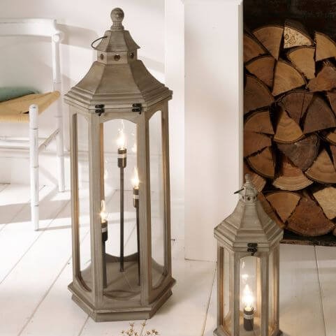 Two antique style lantern table lamps of different sizes, placed on a white wooden floor in front of a white chair and a collection of chopped logs.