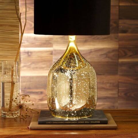 A champagne coloured table lamp with a black velvet lampshade, placed on top of two books on a dark wood surface.