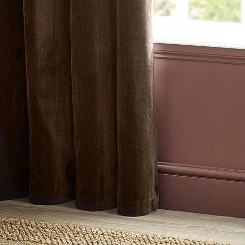 One side of a pair of brown heavy chenille curtains, grazing a wooden floor decorated with a rug, next to a red wall.
