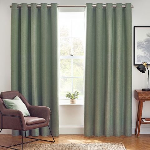 A pair of eucalyptus green thermal blackout curtains, half opened to reveal an outdoor scene, in a white-walled room with a red chair, a woven rug, a wooden side table holding a lamp, a potted plant and a framed piece of wall art.