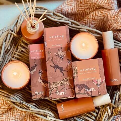 A collection of amber, cinnamon and mandarin scented home fragrance products, including reed diffusers, scented candles and room sprays, arranged in a woven storage basket on top of a rust coloured woven throw.