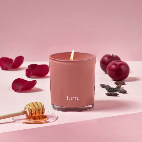 A bergamot, honey, plum and tonka scented glass candle, set on a pink surface with a honey dipper, bergamot petals, plums and tonka beans.