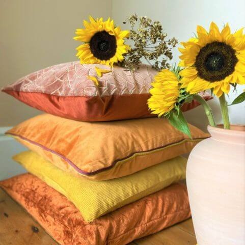 A selection of brick red, orange and yellow scatter cushions with various textured finished, arranged in a single pile alongside three sunflowers in a vase.