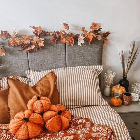 A bedroom decorated in a cosy autumn style, with a burnt orange and white striped duvet cover set, soft novelty pumpkins, brown bed cushions, natural autumn leaves and a decorated side tables. 