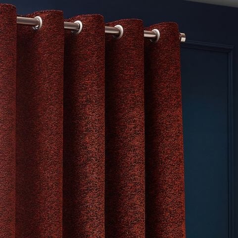 A copper red curtain panel with a stainless steel eyelet heading and a multicoloured mélange design, hung in front of a navy blue wall.