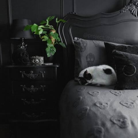 A charcoal Halloween duvet cover set with a tufted skulls design, made on a bed with an ornate black headboard, coordinating black cushions and a decorated side table, while a cat sleeps on the bed.