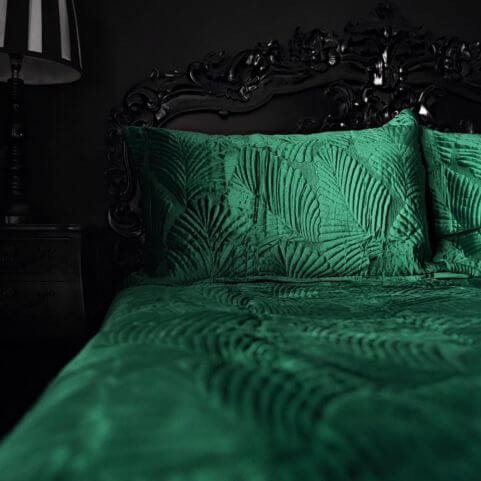 An emerald green duvet cover set with an embroidered quilt velvet design, arranged on a bed with a black headboard, black walls and a black side table holding a lamp.