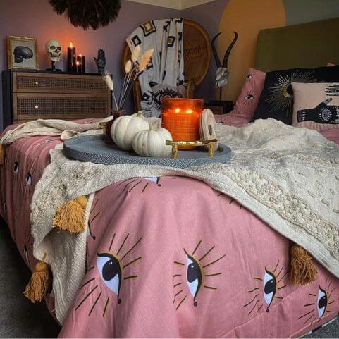 An abstract gothic style bedroom decorated with a pink abstract eye duvet cover set, a tufted boho throw with ochre tassels, abstract bed cushions and a range of gothic accessories.