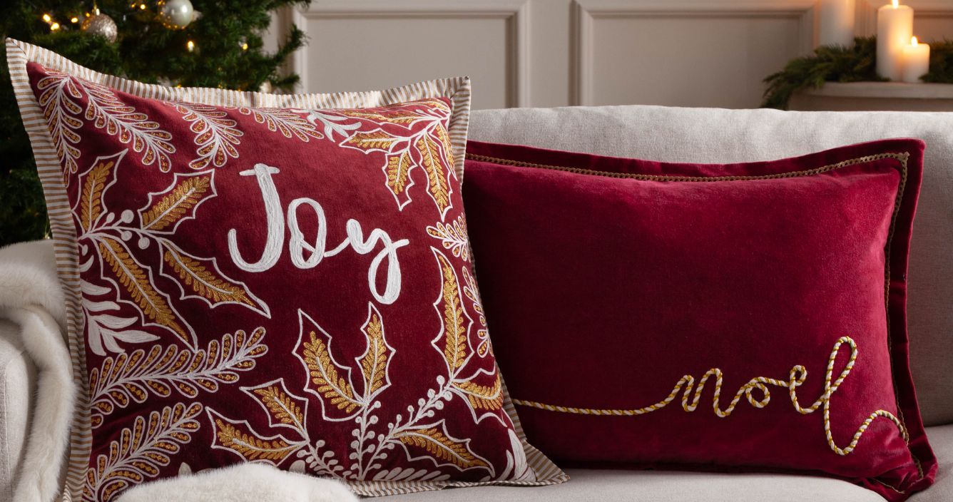 A sofa infront of a Christmas tree, with two Christmas cushion on it saying Joy and Noel