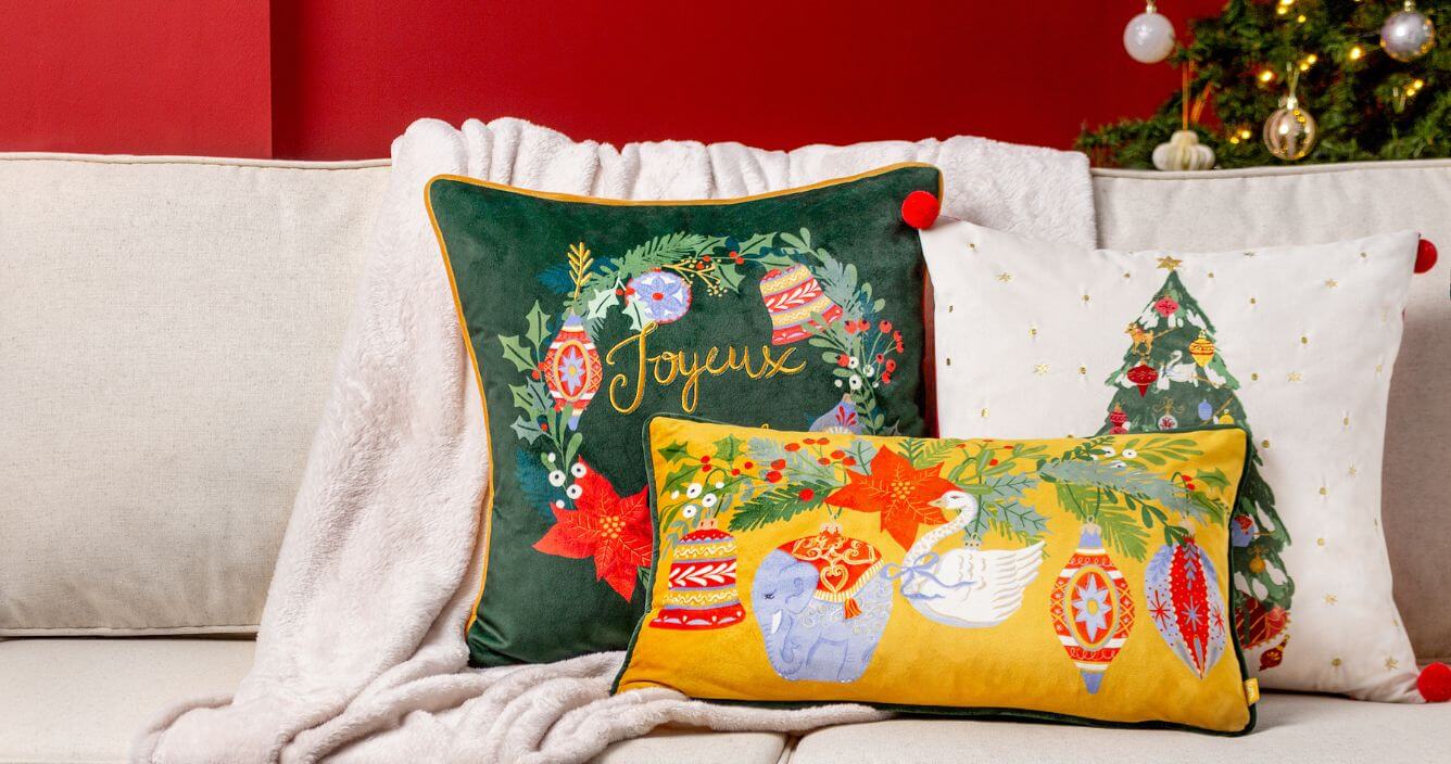 Three green, gold and white Christmas cushions with various festive designs, arranged on a neutral couch with a beige throw.
