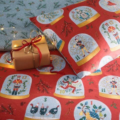 A closeup image of the printed snow globe design on a red Christmas duvet, arranged to reveal the contrasting blue reverse alongside a wrapped Christmas gift.