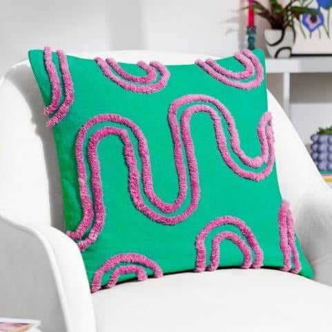 A turquoise cushion with an abstract design of purple cotton tufting, presented on a white chair in front of a neutral background.