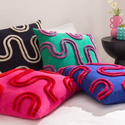 A selection of bright and bold cushions with a matching abstract design of tufted cotton, piled on white bedding in front of a neutral background.