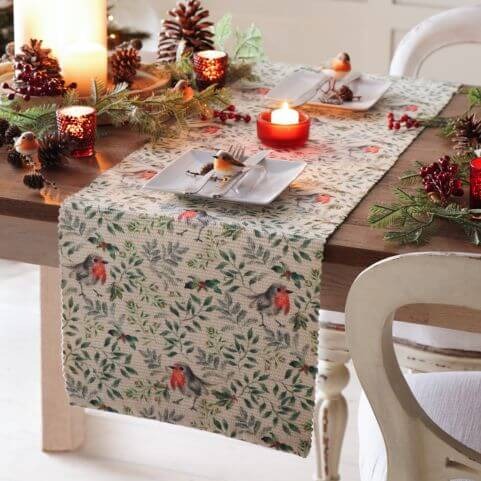 A Christmas table runner with a printed design of robins and festive leaves, presented on a table decorated with pine foliage, candles and novelty robins.
