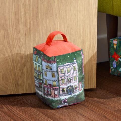 A red and green Christmas door stop with a printed festive town design, laid next to a wooden door on a dark wood floor.