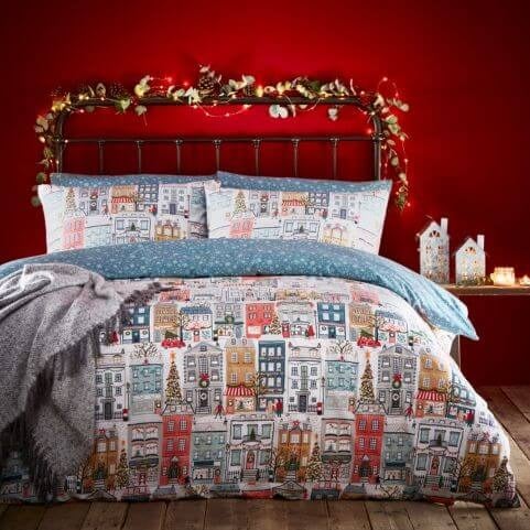 A white Christmas duvet set with a printed design of a festive town, made on a bed in a red room with various seasonal decorations.