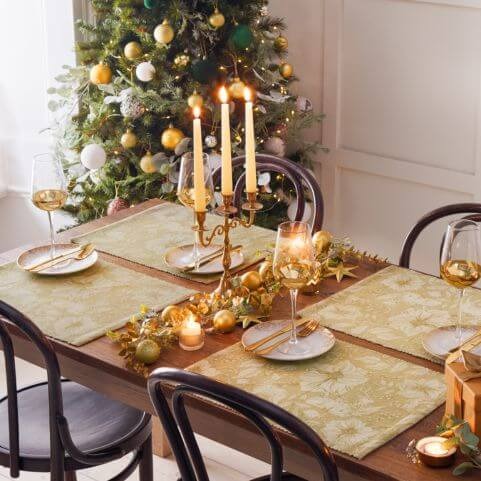 A Christmas table decorated with four gold festive placemats, cutlery, wine glasses, baubles and a candelabra.