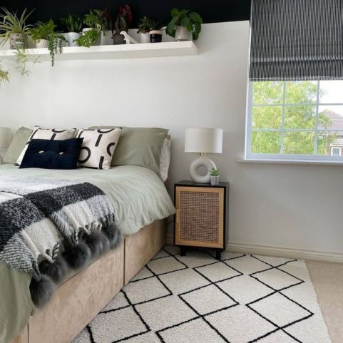 A bright bedroom with white walls. green bedding and monochrome accessories, complete with a collection of houseplants stored on a white shelf above the bed.
