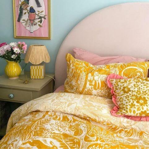 An ochre duvet cover set with a white printed design of winter woodlands, surrounded by a beige side table, flowers in a vase and a yellow table lamp.