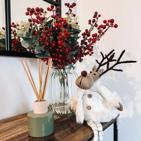 A scented reed diffuser in a green glass bottle, presented on a wooden table alongside a novelty reindeer and red berries in a glass vase.