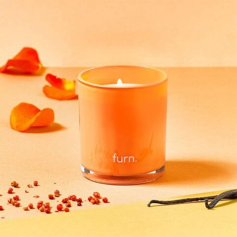 A scented candle in an orange glass holder, placed on an orange surface with vanilla pods, flower petals and other natural elements which make up the scent profile.