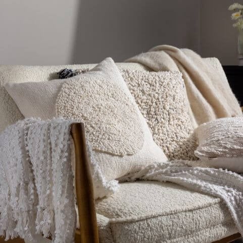 A selection of white cushions and throws with looped yarn bouclé finishes, arranged on a white bouclé sofa in front of a neutral background.