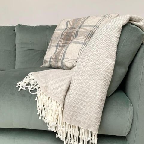 A faux wool beige cushion with a traditional tartan design, placed on a grey sofa with a neutral knitted throw.