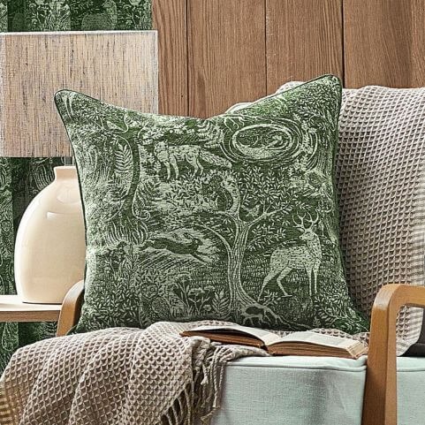 An emerald green cushion with a jacquard design of winter animals among a woodland scene, placed on a pale blue armchair with a waffle weave throw.