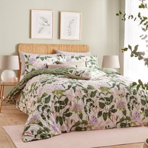 A light green bedroom with a peach and green botanical duvet, two side tables with lamps, wall art and indoor greenery.