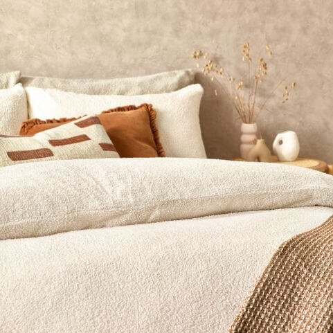 A bed decorated with a white bouclé duvet cover set and rust orange scatter cushions alongside a decorated side table.