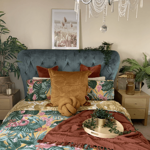 A large bed with a blue velvet headboard, dressed with tropical style bedding, two brown scatter cushions and a red throw.