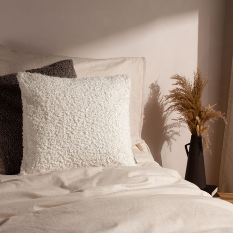 Matching bouclé cushions in differing shades, one white and one grey, propped up in a white bed next to potted foliage.