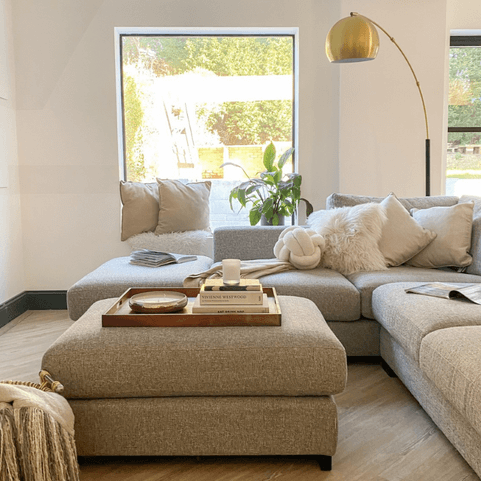 A white living room with a grey sofa, grey ottoman, and a collection of white throw cushions.