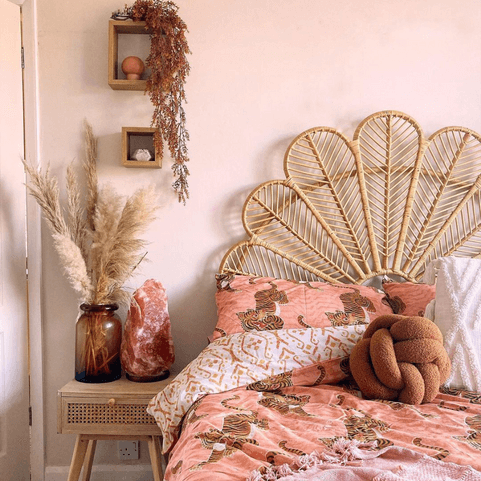 A pink bedroom with a peacock rattan headboard, pink tiger bedding and a ginger boucle knot cushion. There's a brown vase full of pampas grass and a big pink himalayan salt rock lamp on the bedside table.