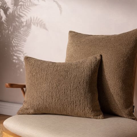 Two matching brown bouclé cushions in differing shapes, displayed together on a grey chair with a wooden frame.
