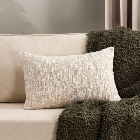 A woven cotton bouclé cushion in a white shade, placed on an off-white sofa with a woolly green bouclé throw.