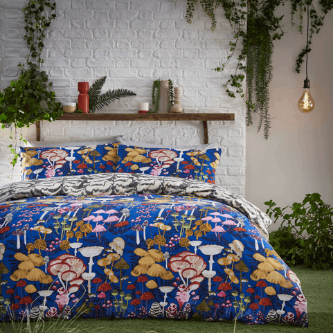 the Amanita bedding set has a cobalt blue background and is decorated with illustrated mushrooms., it is on a bed with a white brick background and hanging green plants